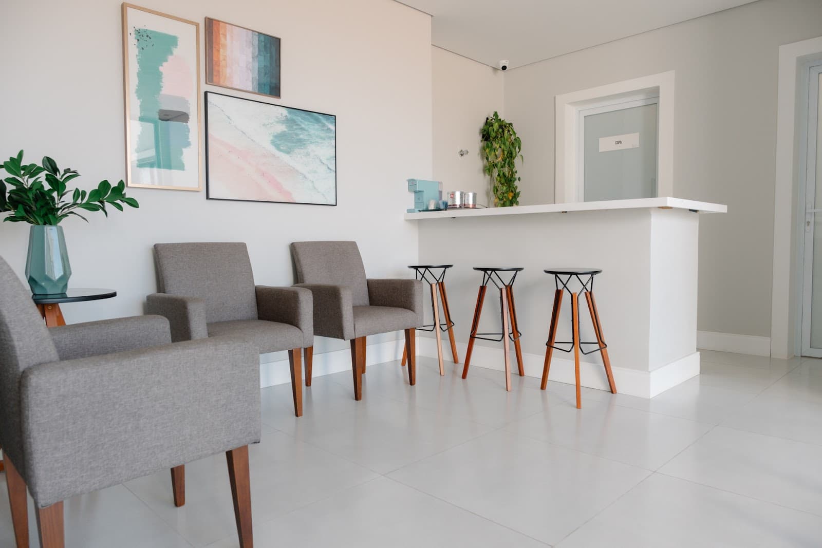 Sala de espera com poltronas cinza, banquetas de madeira, balcão do café e quadros de praia