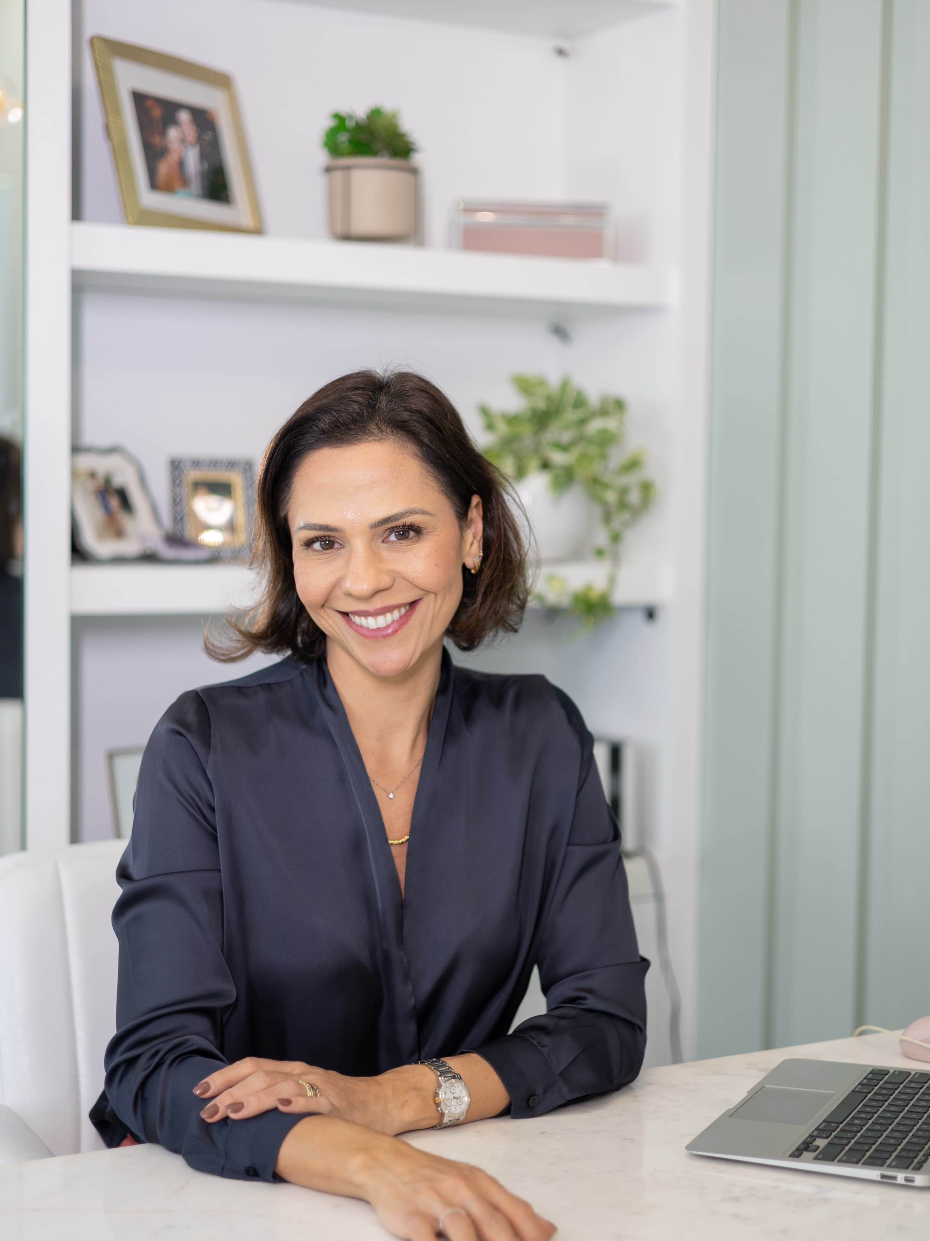 Dra. Ana Carolina sorrindo na mesa do consultório, com a estante clara ao fundo